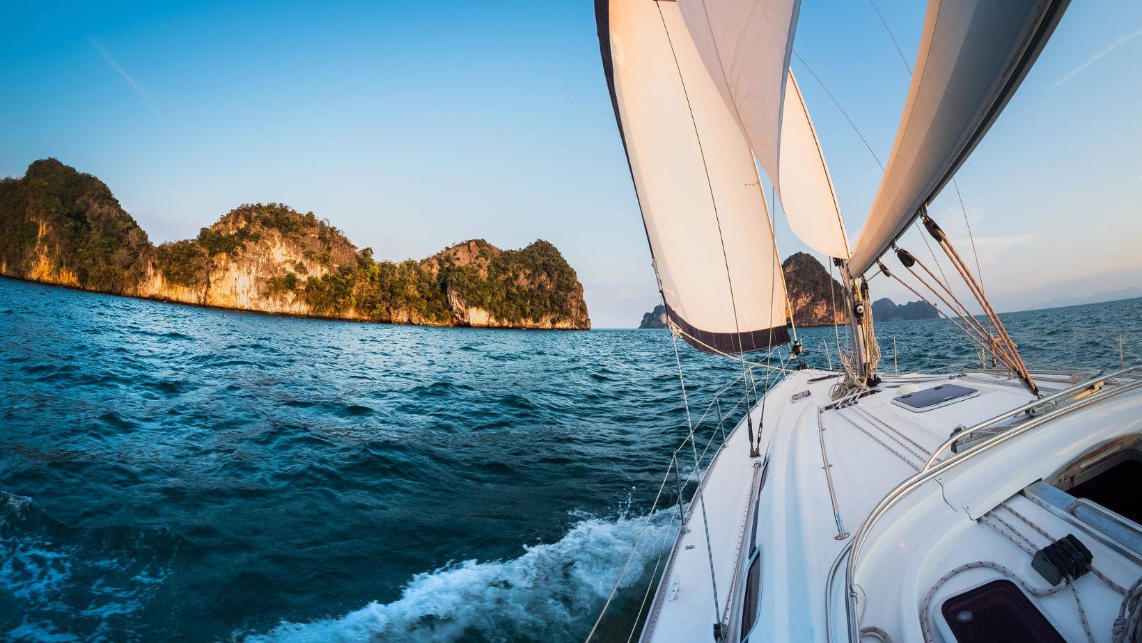 Sailboat navigating turquoise coastal waters past limestone cliffs at sunset – sailing internationally